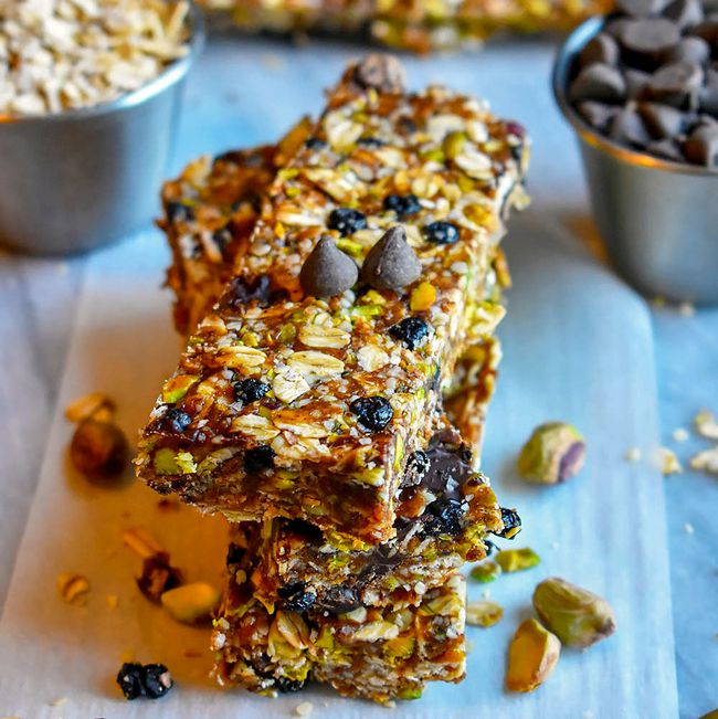 Closeup shot of homemade granola bars on parchment paper, surrounded by pistachios, oats, raisins and chocolate chips