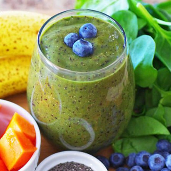 healthy banana smoothie recipes: spinach, blueberries, chia seeds, and bananas frame a glass of a green-colored banana, papaya, and blueberry smoothie, which has three blueberries sitting atop as garnish