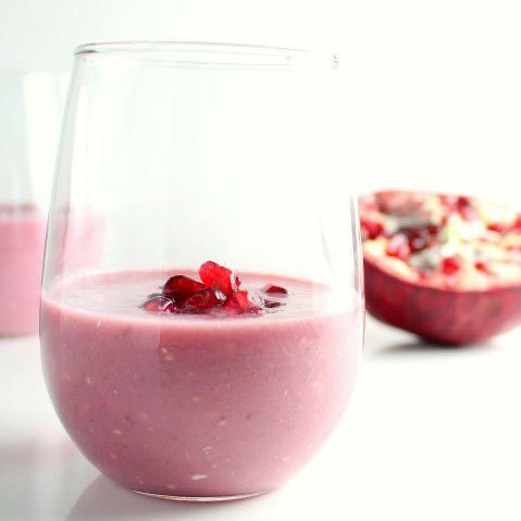 healthy banana smoothie recipes: a stemless wine glass is half full of a pomegranate and banana smoothie with pomegranate arils as a garnish, and another glass and half of a pomegranate are visible in the background